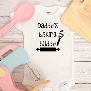 May include: White baby onesie with black text that reads "Daddy's baking buddy" with a rolling pin and whisk graphic.