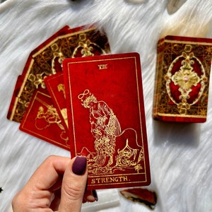May include: A red tarot card with gold illustrations and the word "STRENGTH" at the bottom. The card features a woman with a crown and a lion. Other cards and a box with similar designs are also visible.