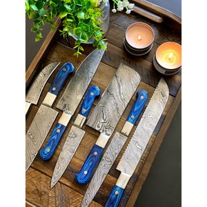Può includere: Set di sette coltelli da cucina blu e argentati con lame a motivi e manici in legno blu su un vassoio in legno.