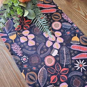 May include: A dark blue table runner with a repeating pattern of pink, orange, and brown stylised flowers and leaves. The pattern is outlined in white.