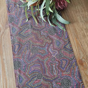 May include: A patterned table runner in a vibrant design of dots in purple, pink, and brown. The design includes circular and swirling patterns. Green leaves and a red flower are placed on top of the runner, adding a natural element.