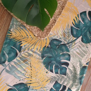 May include: A tropical-themed table runner with a light pink background featuring a repeating pattern of green and yellow leaves. The leaves are stylized and resemble palm fronds and monstera leaves.