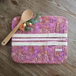 May include: A rectangular kitchen utensil roll with a purple, brown, and white pattern. It has a zip closure and two white straps. A wooden spoon and decorative sprigs are placed on top.