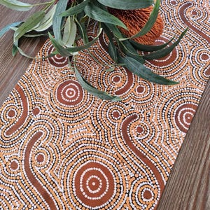 May include: A rectangular table runner with a brown and white dot pattern, featuring a central design of concentric circles and curved lines. Green leaves and a reddish-brown flower are placed on top of the runner, which is set on a wooden surface.