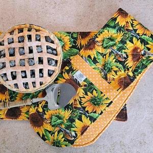 May include: A set of two sunflower-themed kitchen pot holders. The pot holders are made of fabric with a quilted design. One pot holder is a rectangular shape and the other is a square shape. The pot holders are decorated with sunflowers, birds, and polka dots.
