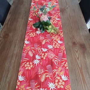May include: Red table runner with a floral pattern featuring white flowers and pink and red buds. The runner is on a wooden table.