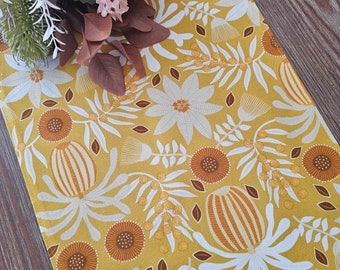 May include: Mustard yellow table runner with a floral pattern in white, orange, and brown. The design features stylized flowers, leaves, and seed pods. A bouquet of faux flowers in shades of pink, brown, and green rests on the runner.