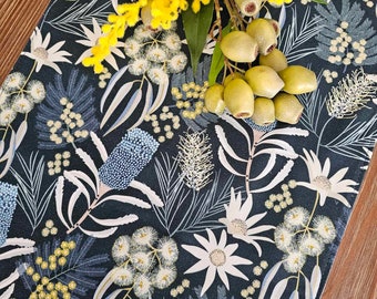 May include: A rectangular fabric table runner featuring a dark navy blue background with a repeating pattern of white and light blue Australian native flora, including banksias and waratahs, topped with a sprig of bright yellow wattle flowers.