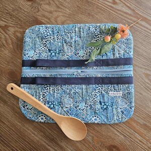 May include: A blue patterned kitchen utensil roll with a zipper closure and navy blue straps. A wooden spoon rests on the wooden surface next to the roll. The roll has a floral accent.