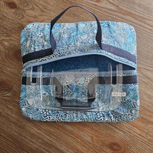 May include: A blue patterned travel bag with a clear, rectangular container inside. The bag has a zip closure and a dark blue handle. The container has a label that reads "Daisy Richi". The bag and container are on a wooden surface.