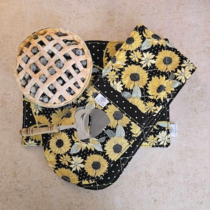 May include: A set of two black and yellow sunflower patterned kitchen pot holders. The pot holders are quilted and have a white polka dot pattern on the back. A wooden cheese knife and a pie filled with blueberries are also in the image.