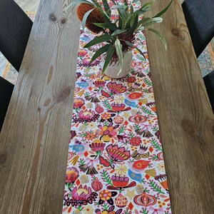 May include: A patterned table runner featuring a floral design in bright colours, including pink, orange, and yellow, set against a white backdrop. A plant in a white pot and a textured flower sit on the runner, placed on a wooden table.