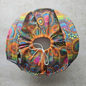 May include: A patterned fabric bowl cover with a drawstring closure. The cover features a vibrant design of orange, yellow, blue, and pink circles and dots. The drawstring is white, and the trim is orange. Designed to fit over a bowl, it measures approximately 25 cm in diameter.