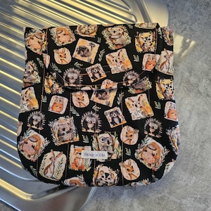 May include: Black fabric bag with a flap closure, patterned with various woodland animal illustrations in shades of brown, orange, and cream. The bag features a repeating pattern of hedgehogs, deer, and koalas. The bag is sitting on a metallic surface.