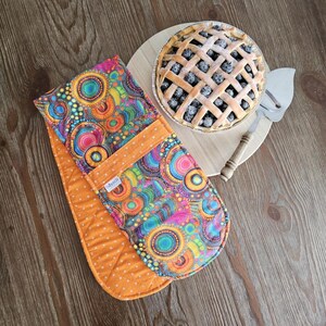 May include: A colorful double oven mitt with a vibrant pattern of circles and dots in shades of blue, green, orange, and pink. Next to it is a blueberry pie on a wooden cutting board with a pie server.