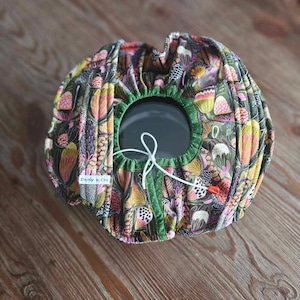 May include: A circular fabric bowl cover featuring a botanical design in green, pink, and yellow hues. It has a green trim and a drawstring. The label "Daisy a. Chi" is visible. The cover is designed to fit over a bowl.