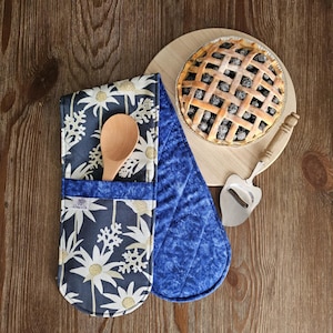 May include: A navy blue oven mitt and pot holder set with a wooden spoon and a blueberry pie. The oven mitt features a white floral pattern. The pie is on a wooden cutting board with a pie server.