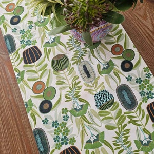 May include: A white table runner with a botanical pattern featuring green leaves and stylized seed pod designs in shades of teal, orange, and black. The runner is placed on a wooden surface, with a vase of greenery in the background.
