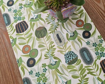 May include: A white table runner featuring a repeating botanical pattern of stylized green leaves, small blue flowers, and rounded seed pods in shades of orange, dark blue, and green. The fabric is laid flat, partially covered by a decorative vase.