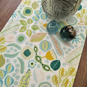 May include: A white table runner with a green and yellow floral pattern. The runner features a variety of leaves, flowers, and seeds. The runner is perfect for adding a touch of nature to your dining table.