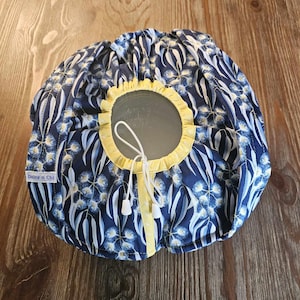 May include: A blue and white fabric bowl cover with a yellow trim and a white drawstring. The cover has a floral pattern with white flowers and blue leaves. The label on the cover reads "Daisy n Chi."