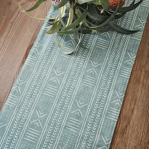 May include: A teal table runner with a white geometric pattern of lines, arrows, and dots. The runner is placed on a wooden surface and adorned with green leaves and orange flowers.