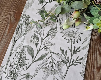 May include: A white table runner with a black and green floral print featuring various Australian native flowers, including proteas, banksias, and gum blossoms.