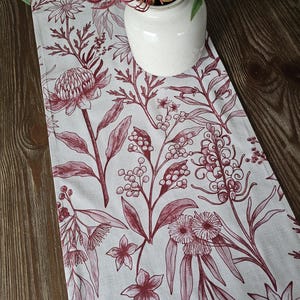 May include: A white table runner with a burgundy floral pattern. The pattern features various flowers, leaves, and stems. The runner is on a wooden table.