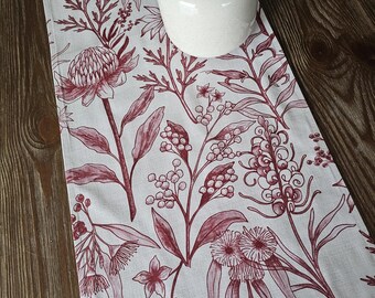 May include: A white table runner with a burgundy floral pattern. The pattern features various flowers, leaves, and stems. The runner is on a wooden table.