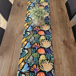 May include: A patterned table runner featuring a black backdrop and a bright floral design. The pattern incorporates orange, yellow, blue, and purple flowers and foliage. A decorative plant in a silver pot sits on the runner, which is placed on a wooden table.