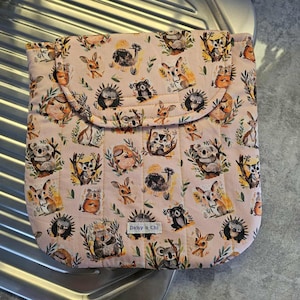 May include: A pink quilted fabric case featuring a repeating pattern of various animals, including koalas, hedgehogs, and deer, amidst leafy branches. The case has a flap closure and a label that reads "Daisy n Chi".