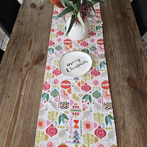 May include: A rectangular table runner featuring a white background with a repeating pattern of colourful Christmas ornaments, stylised trees, gift boxes, and kangaroos. A white ceramic dish with the text merry Christmas sits on the runner.