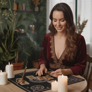 May include: A woman in a burgundy dress with gold embroidery sits at a round table, reading tarot cards. Lit candles and an incense burner are on the table. A black mat with a circular design is under the cards.