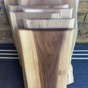 May include: A stack of wooden cutting boards with natural edges. The boards vary in size and shape, showcasing the wood's grain and color variations, ranging from light tan to rich brown. The boards are displayed against a brick wall and a blue and white striped surface.