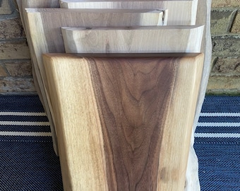 Puede incluir: Un conjunto de tablas de cortar de madera con bordes naturales. Las tablas varían en tamaño y forma, mostrando las vetas y variaciones de color de la madera, que van desde el beige claro hasta el marrón oscuro. Las tablas se exhiben contra una pared de ladrillo y una superficie a rayas azules y blancas.