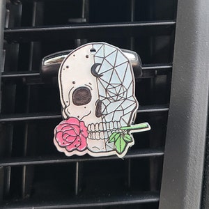 May include: A decorative car air vent clip featuring a skull design with a geometric pattern on one half and a pink rose in its mouth. The clip is silver and the skull is white with black accents.