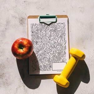 Pu&ograve; includere: Una pagina da colorare in bianco e nero con un disegno floreale su una clipboard. Una mela rossa e un manubrio giallo sono su una superficie grigia.