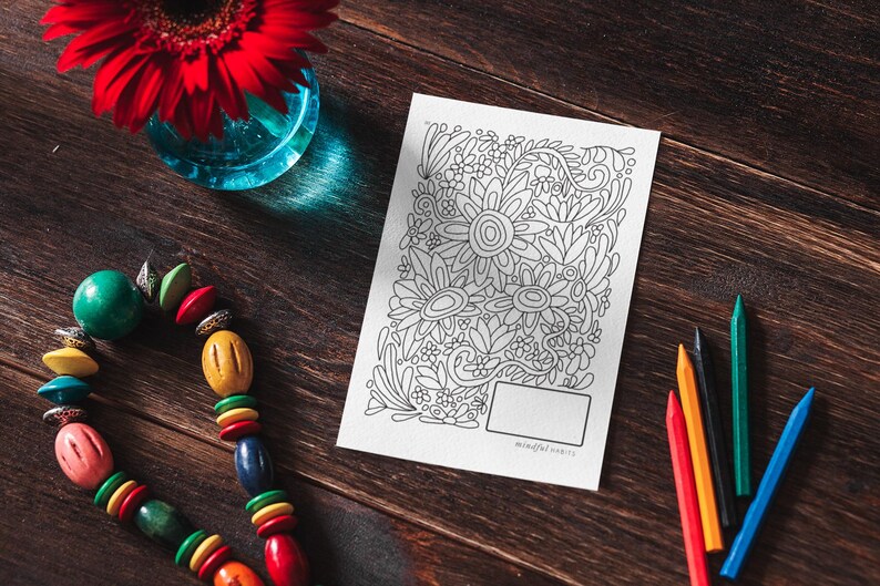 Pu&ograve; includere: Una pagina da colorare in bianco e nero con un disegno floreale. La pagina &egrave; su un tavolo di legno con un fiore rosso, una collana di perline colorate e matite colorate.