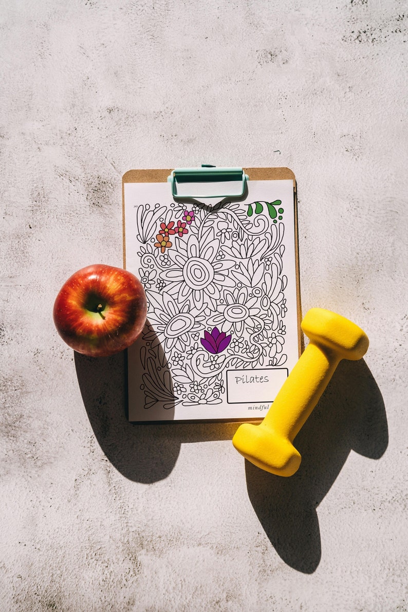 Pu&ograve; includere: Una cartellina bianca con un disegno floreale in bianco e nero e la parola "Pilates" scritta su di essa. Nell'immagine sono presenti anche un manubrio giallo e una mela rossa.