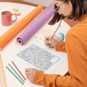 Pu&ograve; includere: Una persona sta colorando un disegno floreale dettagliato in bianco e nero su un foglio di carta con una matita. La persona &egrave; seduta a un tavolo con un righello, matite e due rotoli di carta da regalo.