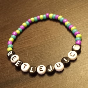 Op de afbeelding: Een kralenarmband met het woord "BEETLEJUICE" gespeld in zwarte en witte letterkralen. De armband is gemaakt met groene, paarse en blauwe kralen.