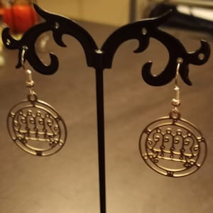 May include: A pair of gold-toned earrings with a circular design featuring a stylized image of a crown. The earrings are hanging from a black earring stand.
