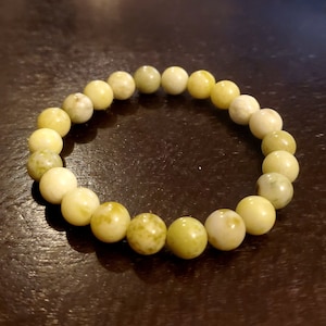 Op de afbeelding: Een armband bestaande uit ronde, lichtgeelgroene edelsteen kralen. De armband is elastisch en aaneengeregen, waarbij elke kraal ongeveer 1 cm in diameter is. De kralen hebben een gevlekt uiterlijk met kleurvariaties.