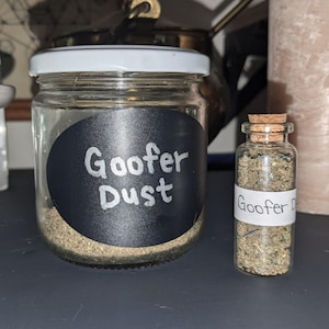 May include: Two glass jars with cork lids filled with a brown powder. The larger jar has a black label with white text that reads "Gooofer Dust". The smaller jar has a white label with black text that reads "Gooofer Dust".