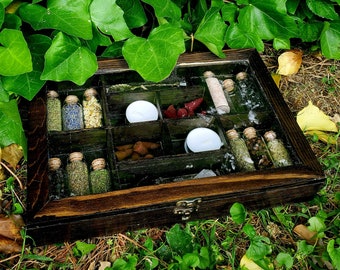 May include: A wooden box with a glass lid containing small glass bottles filled with dried herbs and flowers. The box has a latch and is surrounded by green foliage.