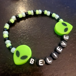 May include: A green and black beaded bracelet with the word "BELIEVE" spelled out in white letters on black beads. The bracelet also features two green alien head charms.