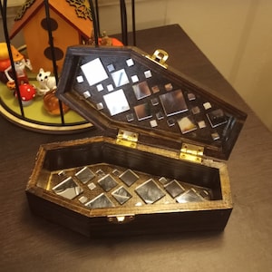 Könnte beinhalten: Eine Holzkiste in Sargform mit einem Scharnierdeckel. Der Deckel und das Innere sind mit kleinen Spiegelquadraten verziert.