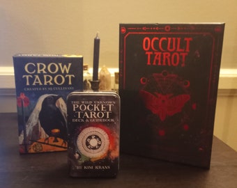 May include: Three tarot card decks, two with black backgrounds and red lettering. The decks are titled "Crow Tarot", "Occult Tarot", and "The Wild Unknown Pocket Tarot".