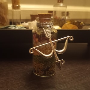 May include: A clear glass bottle with a cork stopper filled with dried herbs, flowers, and seeds. A silver bow and arrow charm is attached to the bottle.