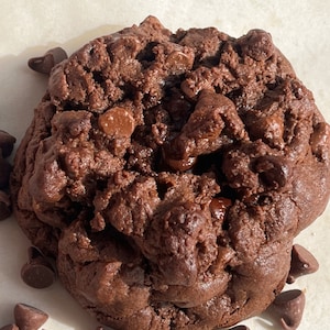 Puede incluir: Una galleta grande de chocolate con chispas de chocolate oscuro, con una textura masticable y una generosa cantidad de chispas de chocolate. La galleta está rodeada de chispas de chocolate más pequeñas.